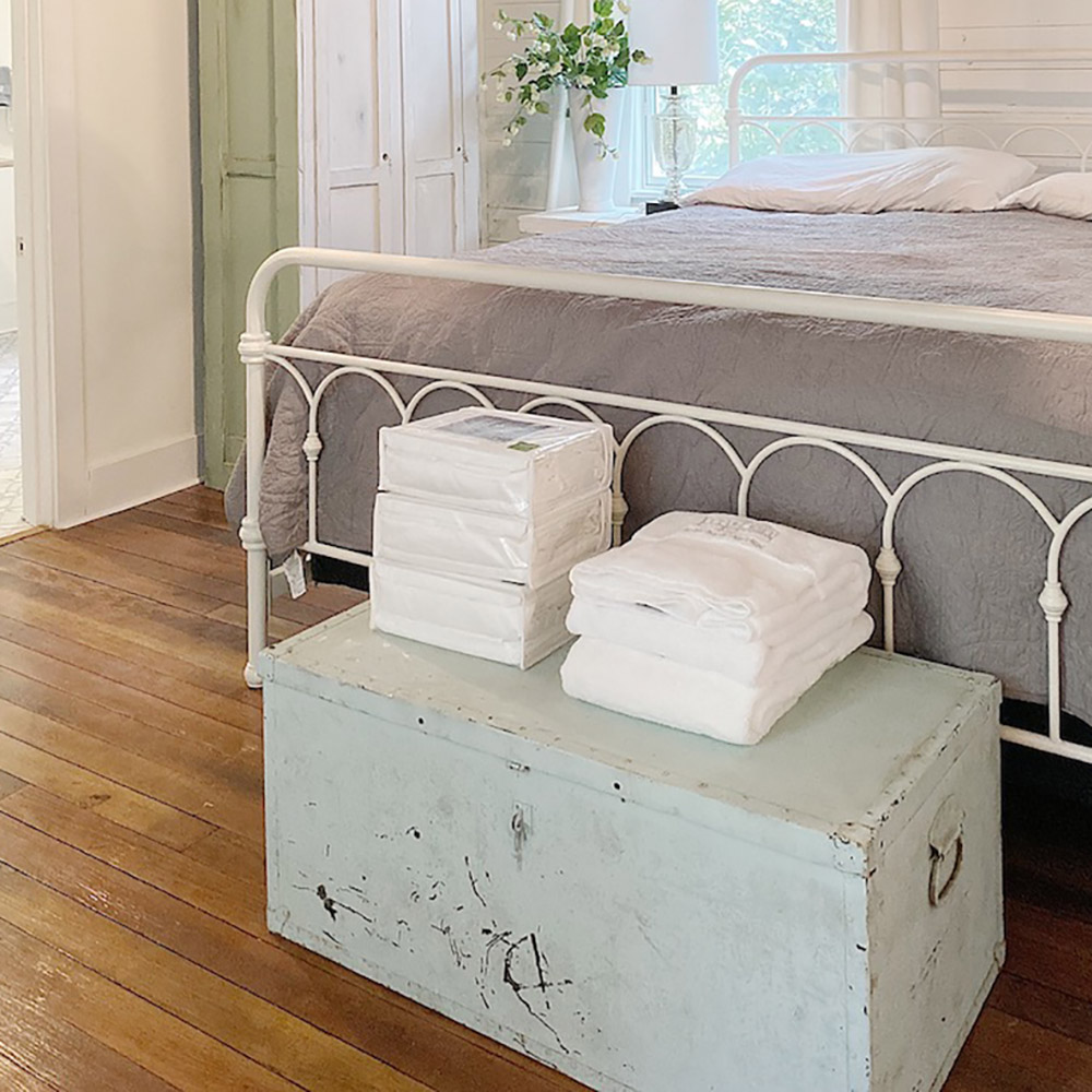 A stack of sheets and towels sitting on a light-colored chest at the foot of a bed.