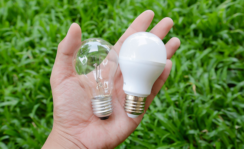 A person holding an incandescent light bulb on the left and an LED light bulb on the right.