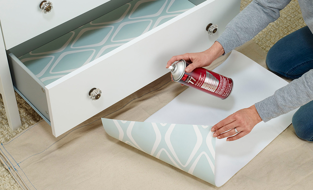 A person uses spray adhesive on a piece of drawer liner paper. 