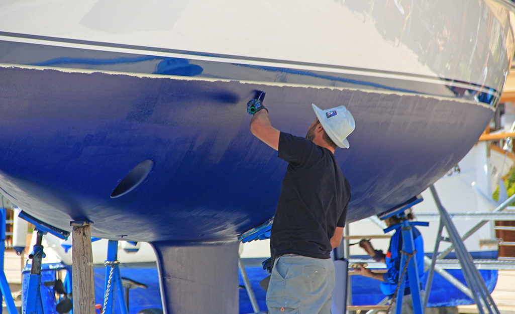 A man wearing a t-shirt, shorts and a hat applies blue paint to the bottom of a dry-docked boat.