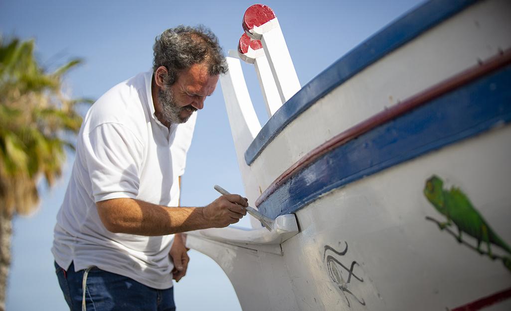 A bearded man uses a brush to touch up the paint on a white boat with blue and red trim.