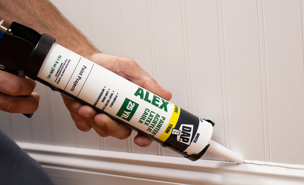 Person using a caulk gun with a latex caulk cartridge on wood trim