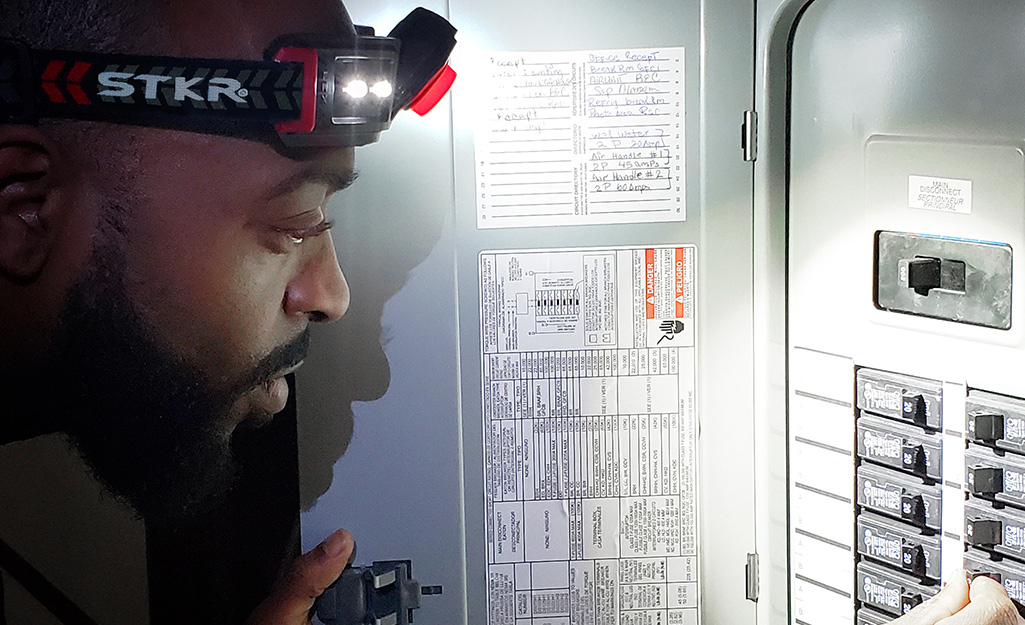 A person wearing a headlamp adjusts a circuit breaker in an electrical panel.
