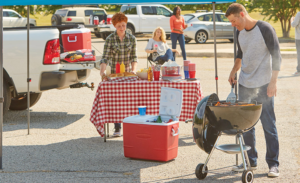 A man cooks food on a grill while a woman sets up a table with a checked cloth next to a red cooler at a tailgate.