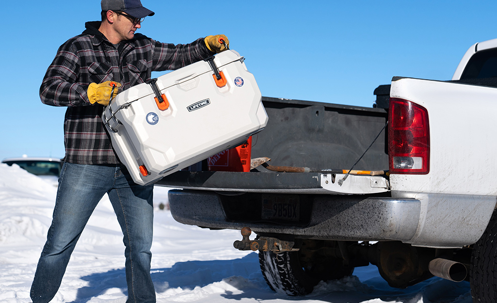 A man puts a white cooler into the back of a white truck parked on snowy ground.