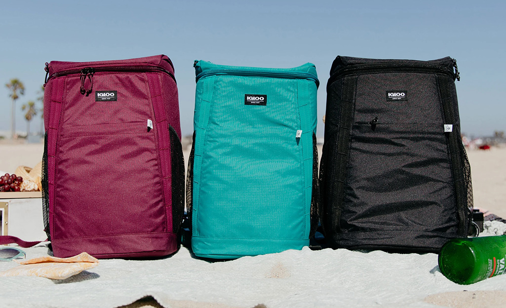 A maroon backpack cooler sits next to an aqua backpack cooler and a black backpack cooler on a beach.