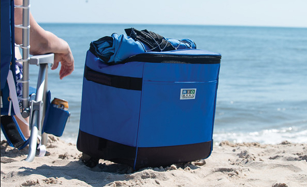 A blue cooler stands next to a person in a beach chair sitting by the ocean.