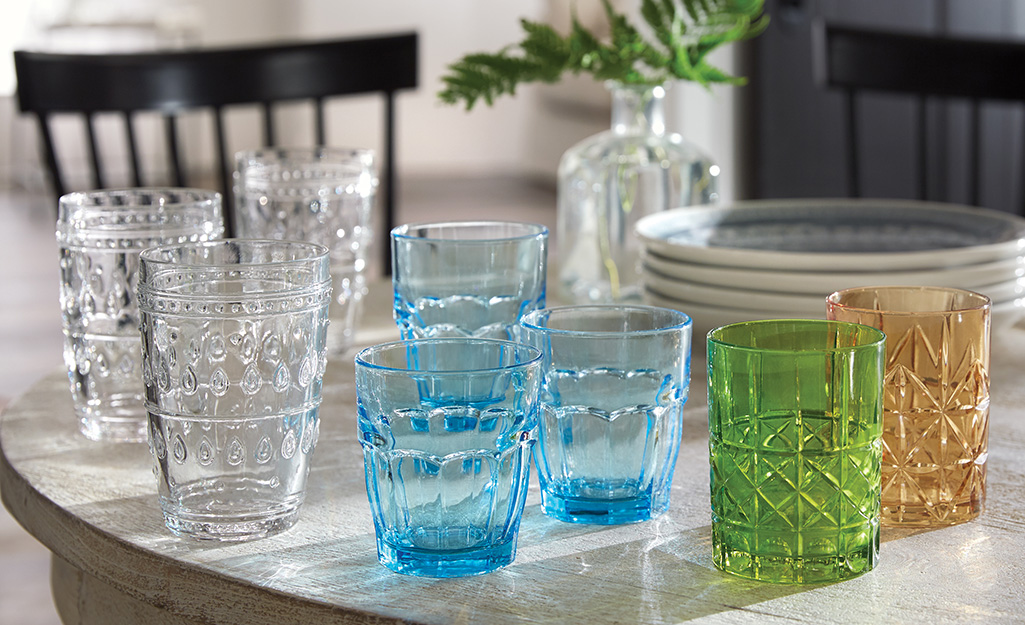 Cut-glass drinking glasses in several solid colors sit on top of a table in front a stack of plates.