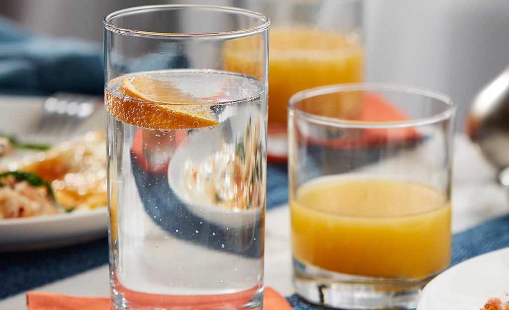 A tall glass with water in it sits next to a shorter glass of orange juice next to two plates on a table.