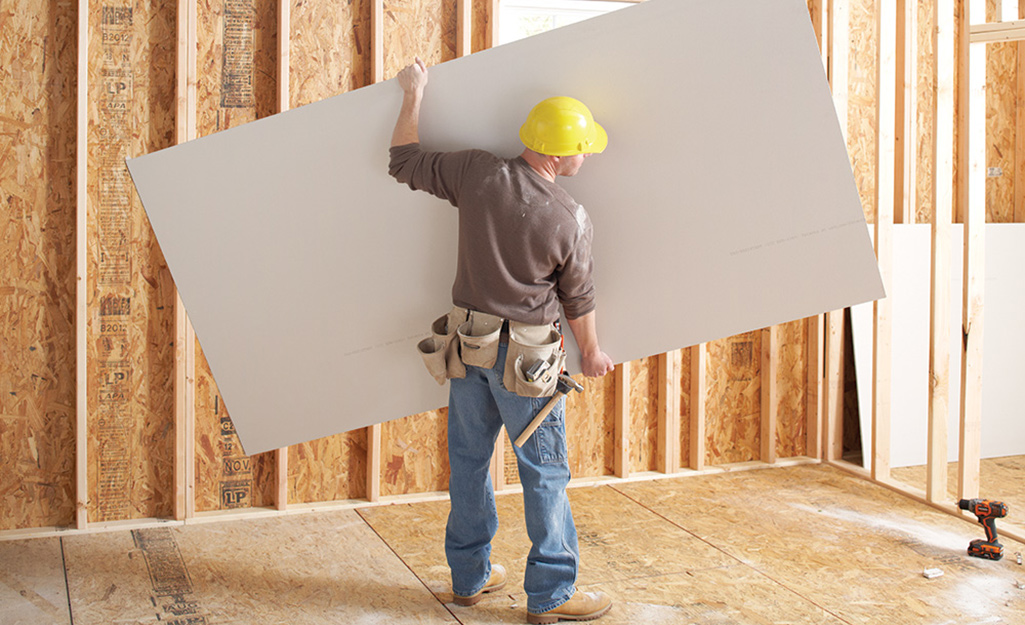 A man holding a large sheet of drywall in an unfinished space.