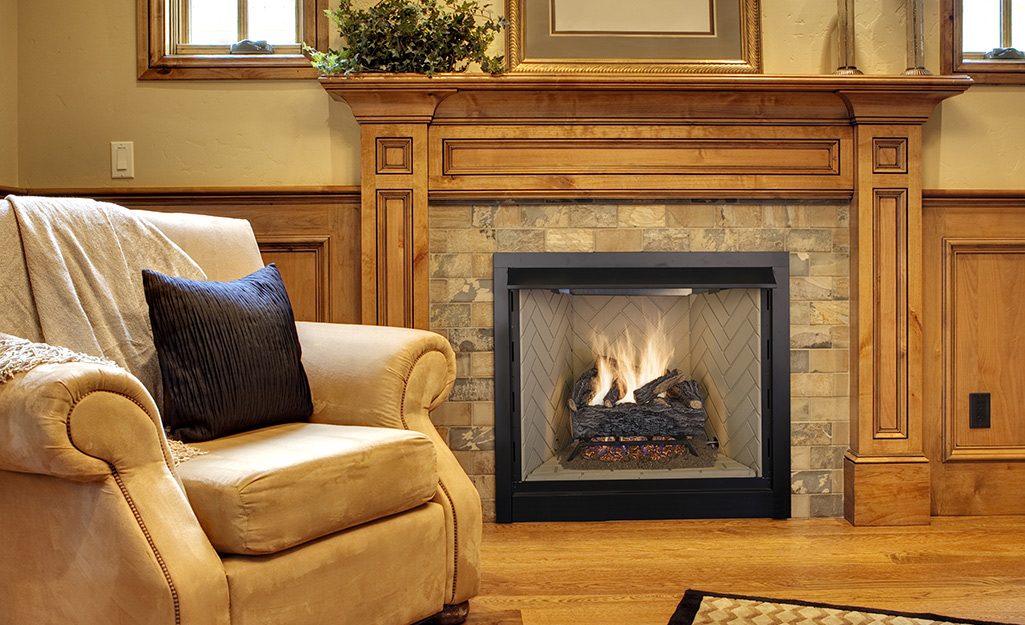An upholstered chair sits near a lit gas fireplace with a stone surround and a wooden mantel that matches the wall paneling.