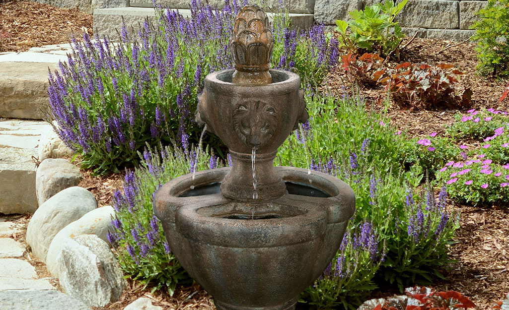 A tiered water fountain in a flower bed next to a walkway.
