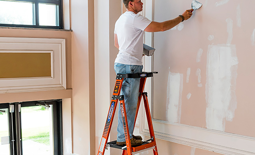 A man patching a wall while standing safely on a platform ladder.