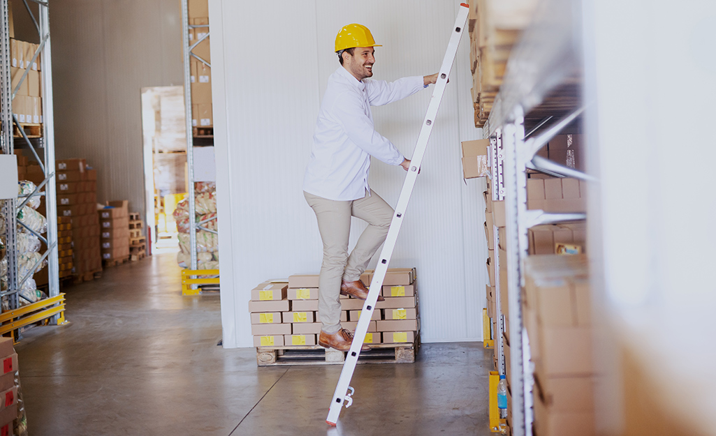 A man climbing a straight ladder leaned against shelves in a warehouse.