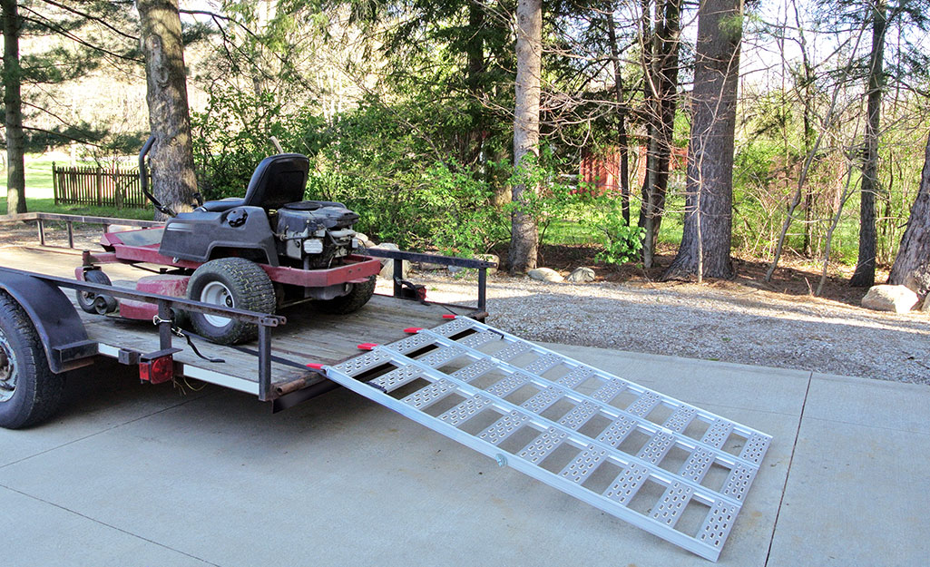 A loading ramp connects to a flatbed trailer with a riding lawn mower in it.