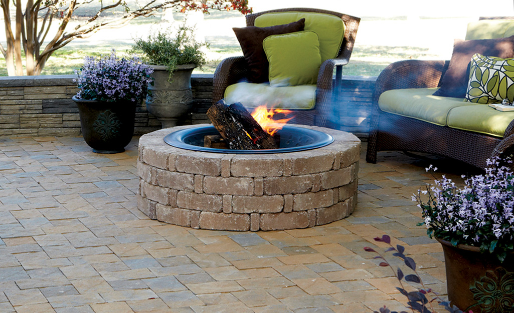 A firepit and patio made of paving stones with a chair and, love seat and pots of blooming flowers.