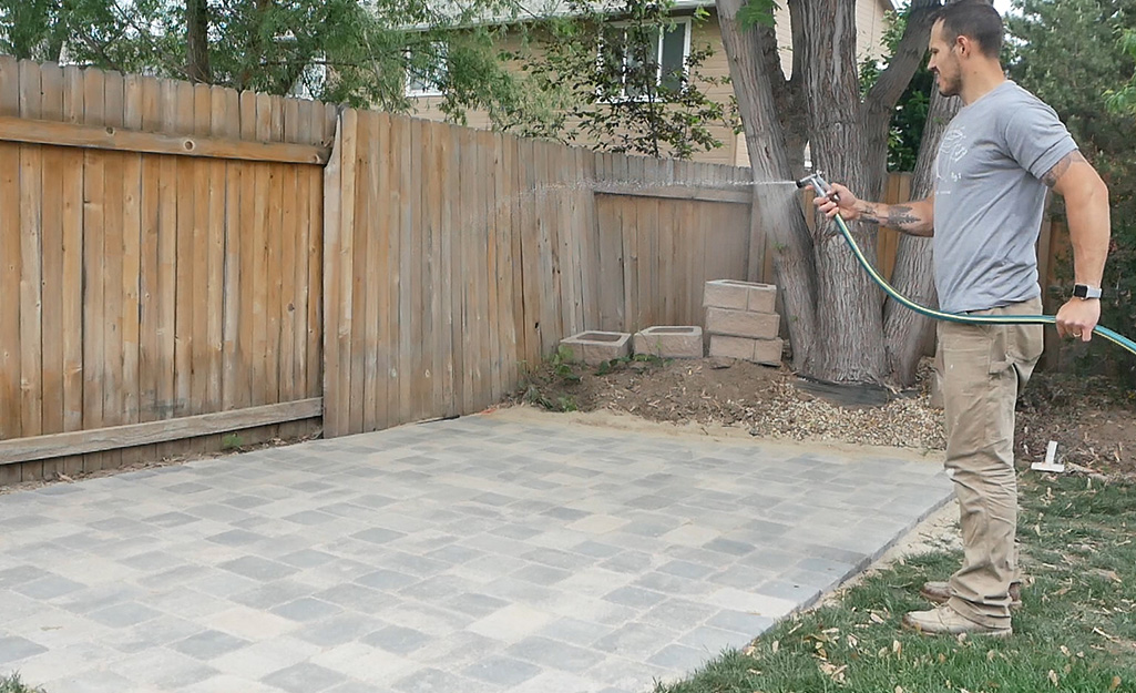 A man using a hose and spray nozzle to clean off a paver patio.