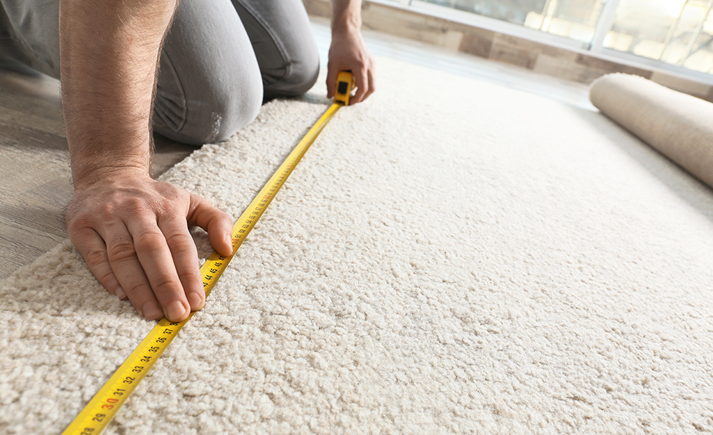 A person kneels on the floor to measure a rug.