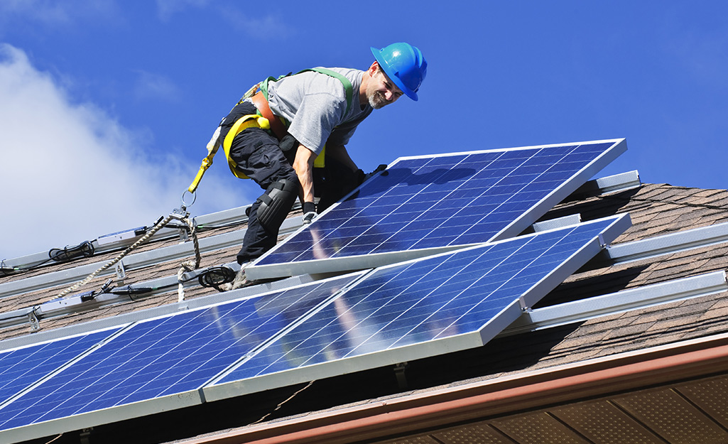 A man wearing a blue hard hat and a yellow safety harness bends to lift a solar panel on the roof of a house.