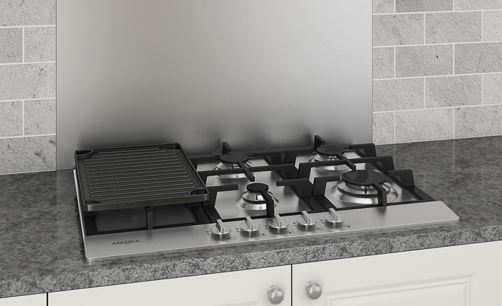 A gas stovetop has a grill pan bridge accessory to the left of four burners.