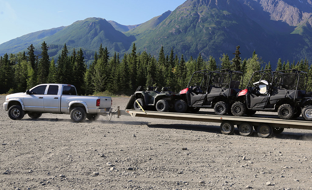 A white pickup truck with a flatbed trailer loaded with ATVs parks in front of trees and mountains.