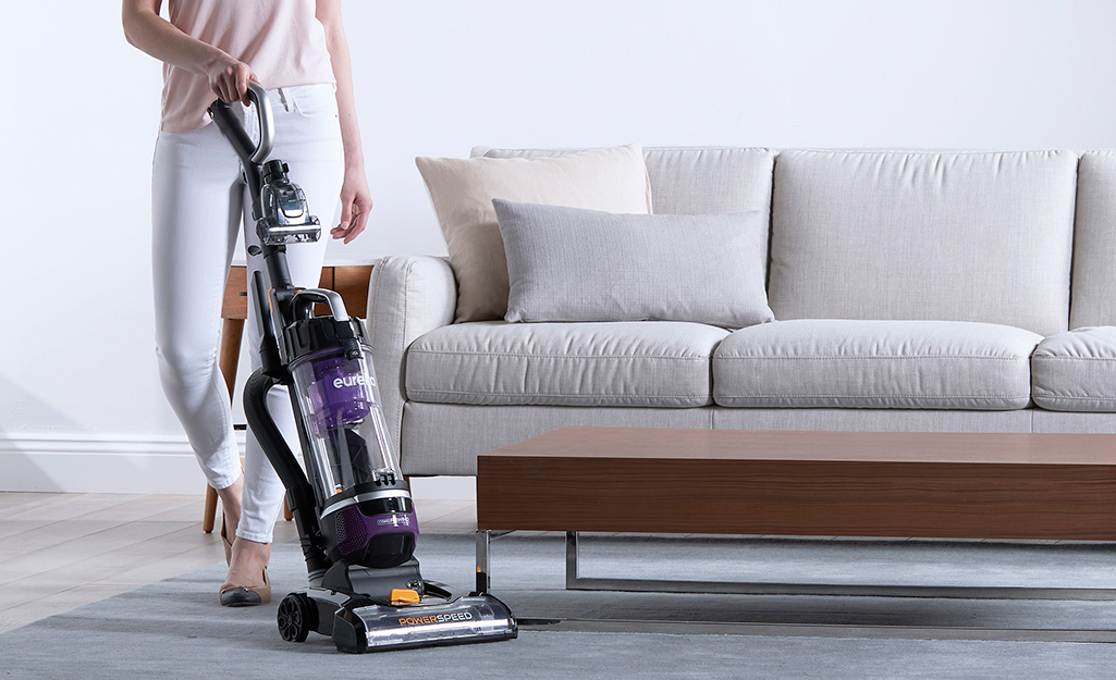 A person vacuuming in front of a sofa with an upright vacuum cleaner.