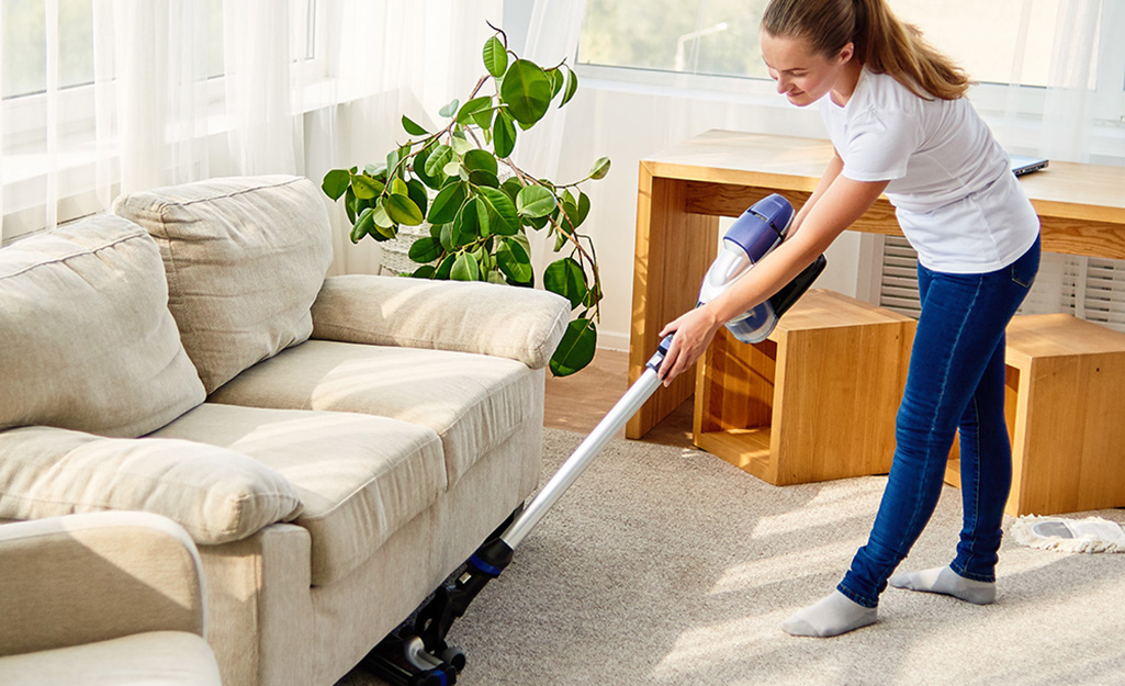 A person pushing a vacuum cleaner along the carpet under a sofa.