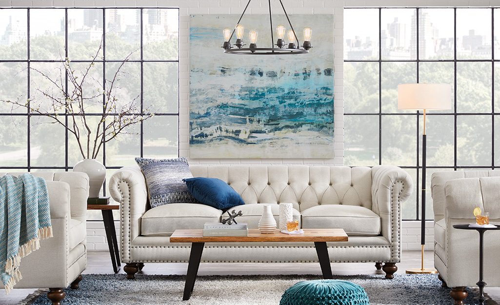 Floor to ceiling windows act as a backdrop for a tufted white couch and two matching chairs in a white living room with blue accents.