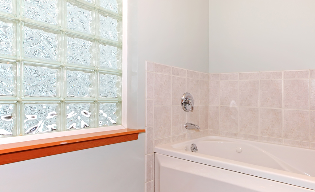 Light comes through a glass block window next to a bathtub in the corner of a bathroom.