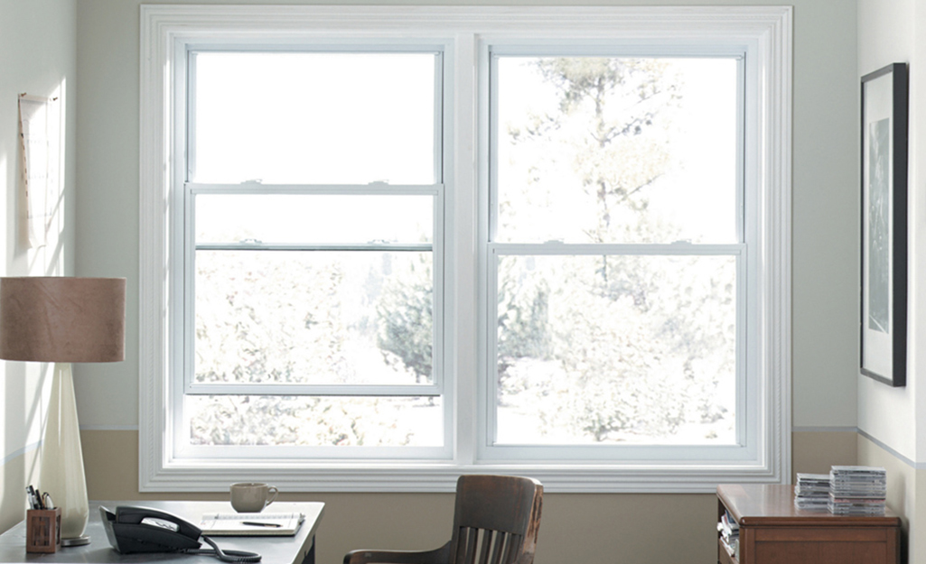 A desk and chair sit in front of a window in a vinyl frame in a small home office.