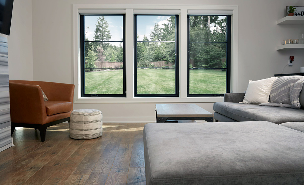 A sectional couch and leather chair sit in front of three casement windows with a view of a lawn.