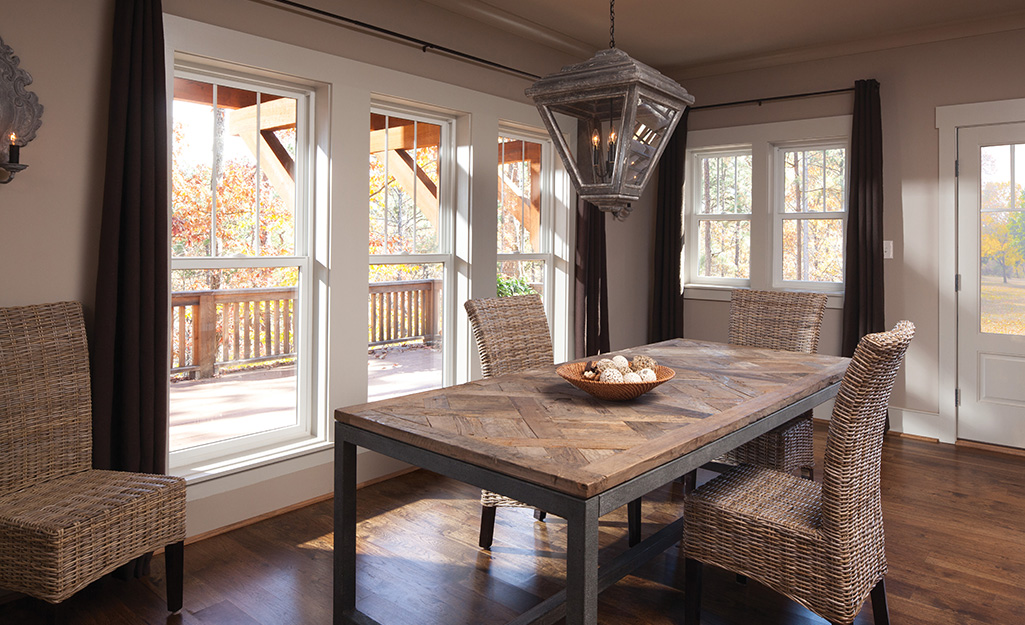 A dining room table and four wicker chairs stand in front of single hung windows that open onto a deck.
