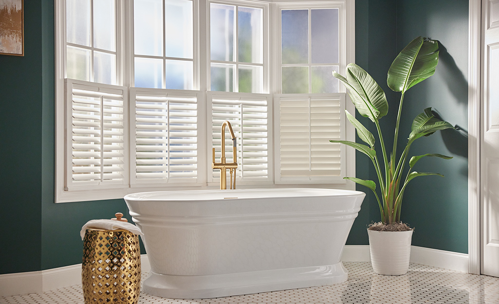A freestanding bathtub sits in front of a bay window that has white shutters over its bottom half.