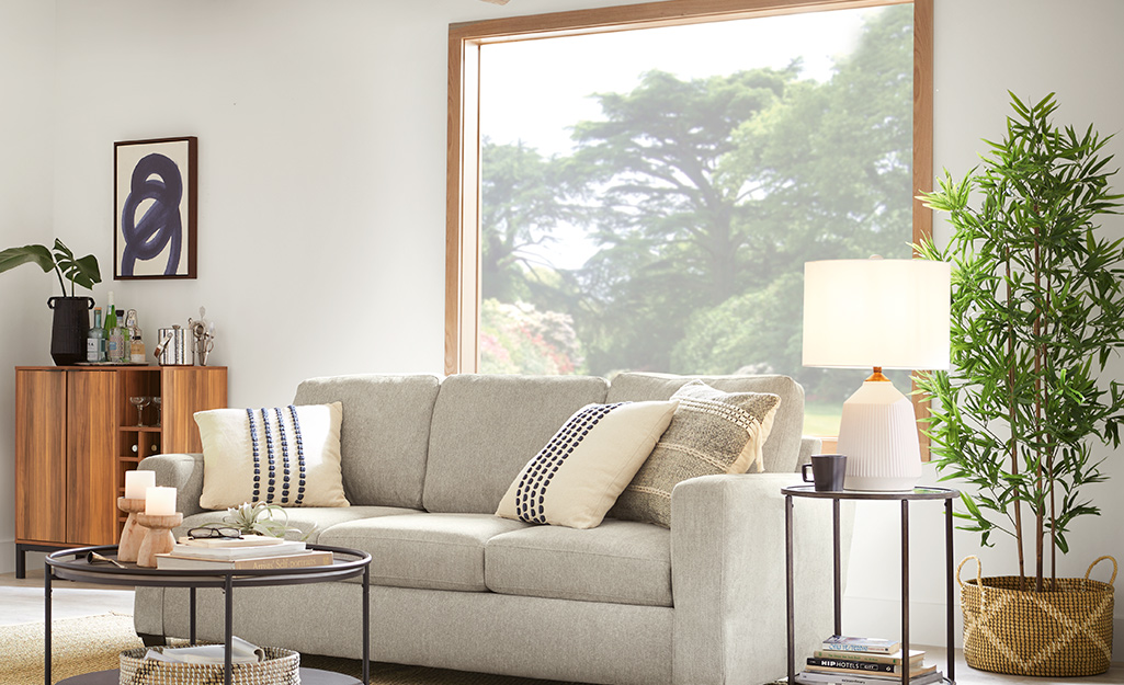 A beige couch sits in front of a picture window that offers a view of a yard and several mature trees.