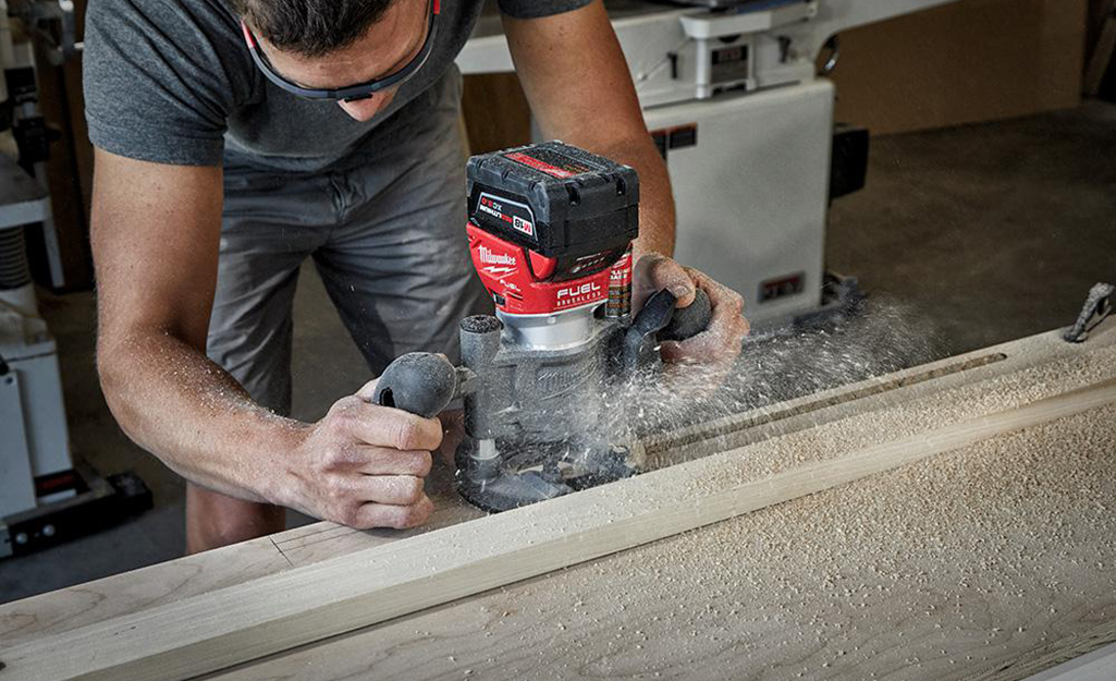 A man wearing safety glasses uses a wood router to a trench in a piece of wood. Sawdust flies off the tool.