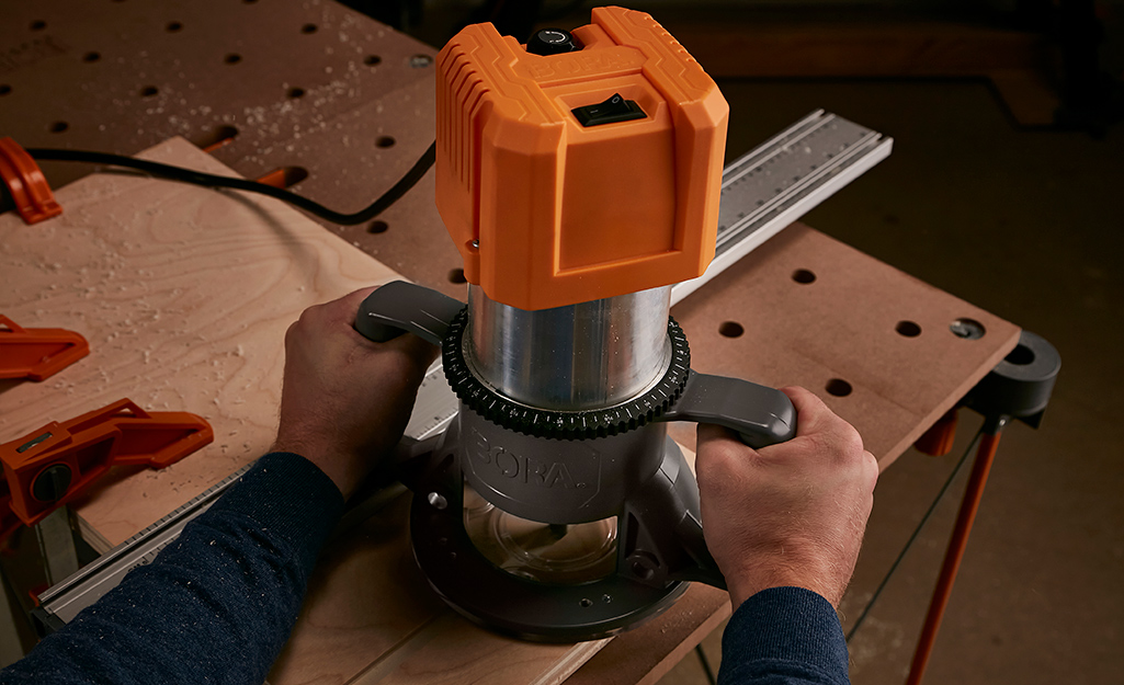 Two hands hold a wood router as a cut is made at the edge of a wood project in a workshop.