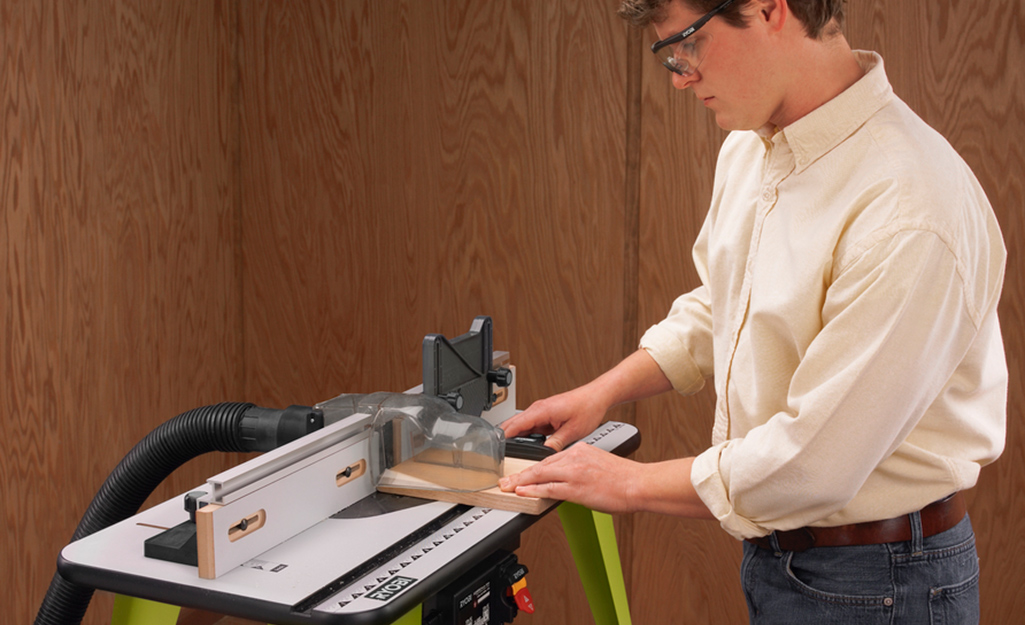A man wearing safety glasses stands at a wood router table to use a wood router tool in a wood-paneled room.  