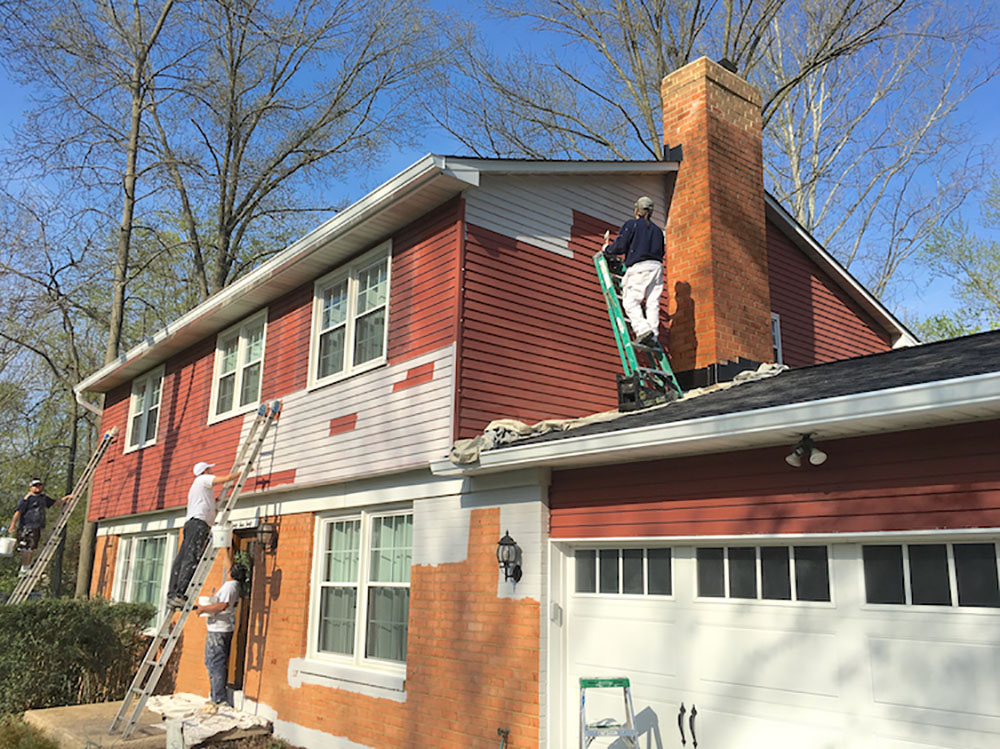 A group of men paint the exterior of a home with red siding and orange brick.