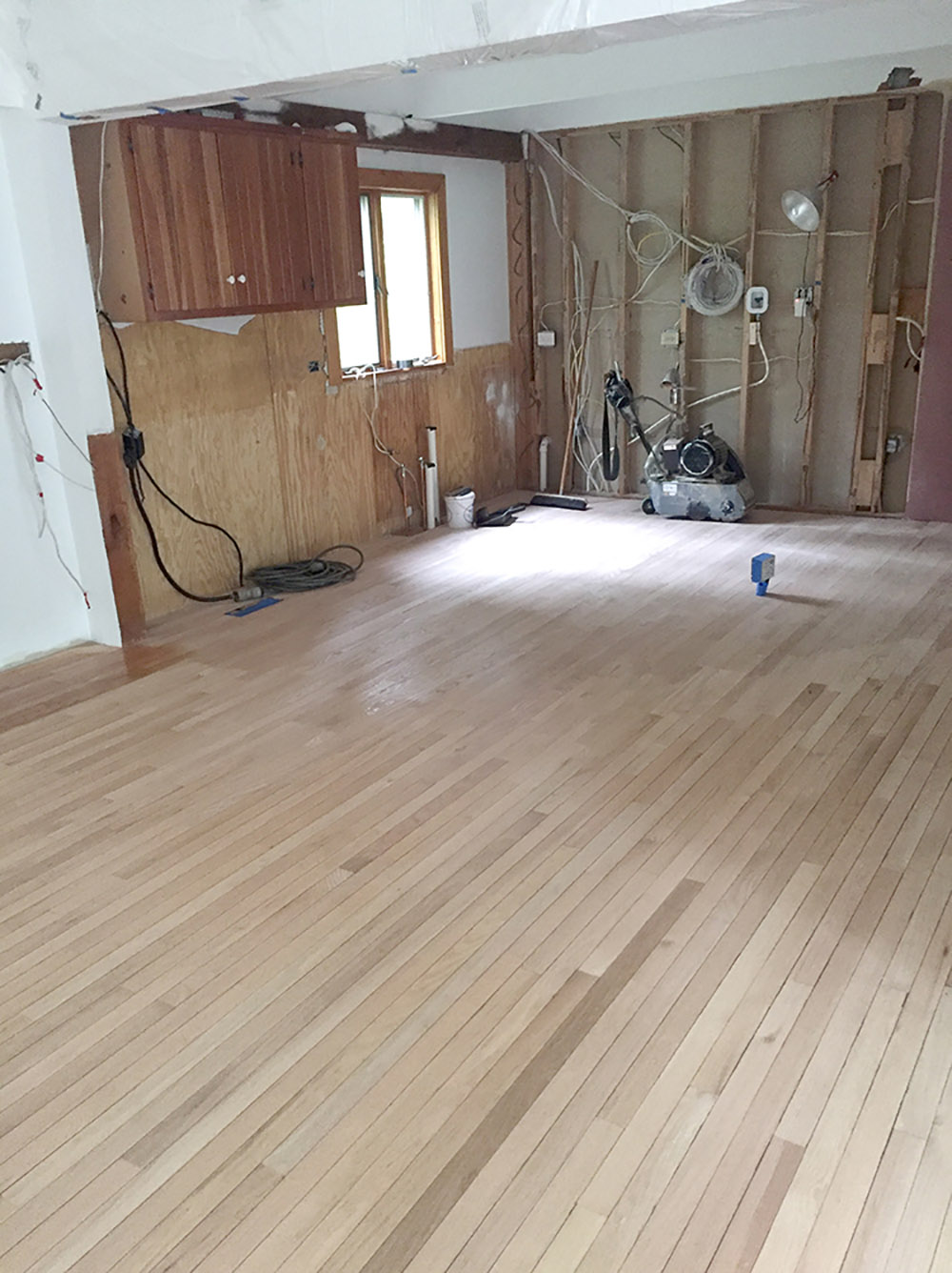 An open and empty kitchen down to the studs with new unfinished hardwood floors.