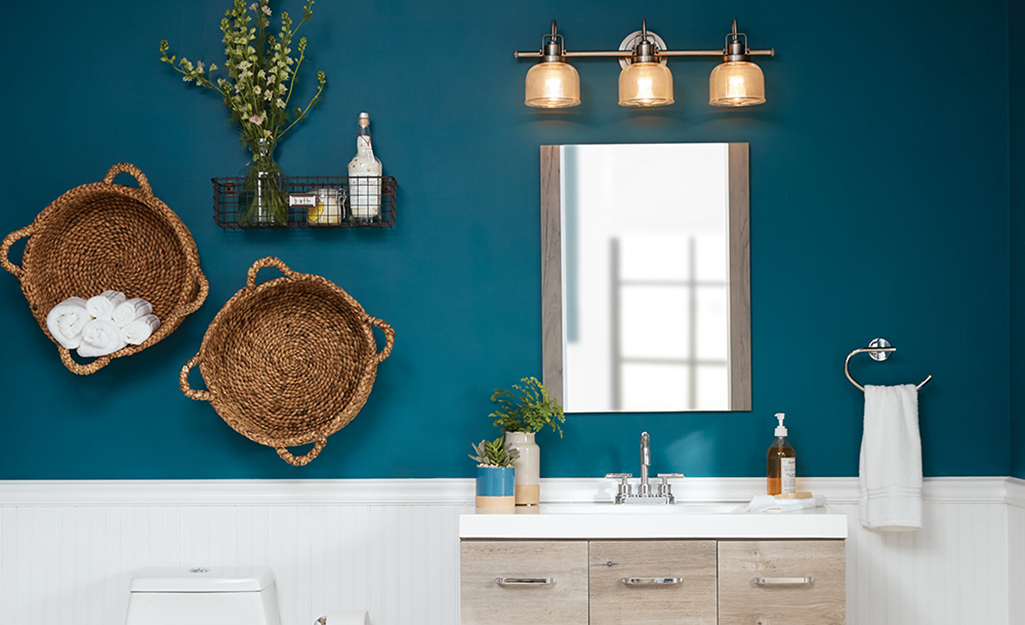 A vanity light bar with three glass sconces hangs above a mirror next to two round baskets on a teal bathroom wall.