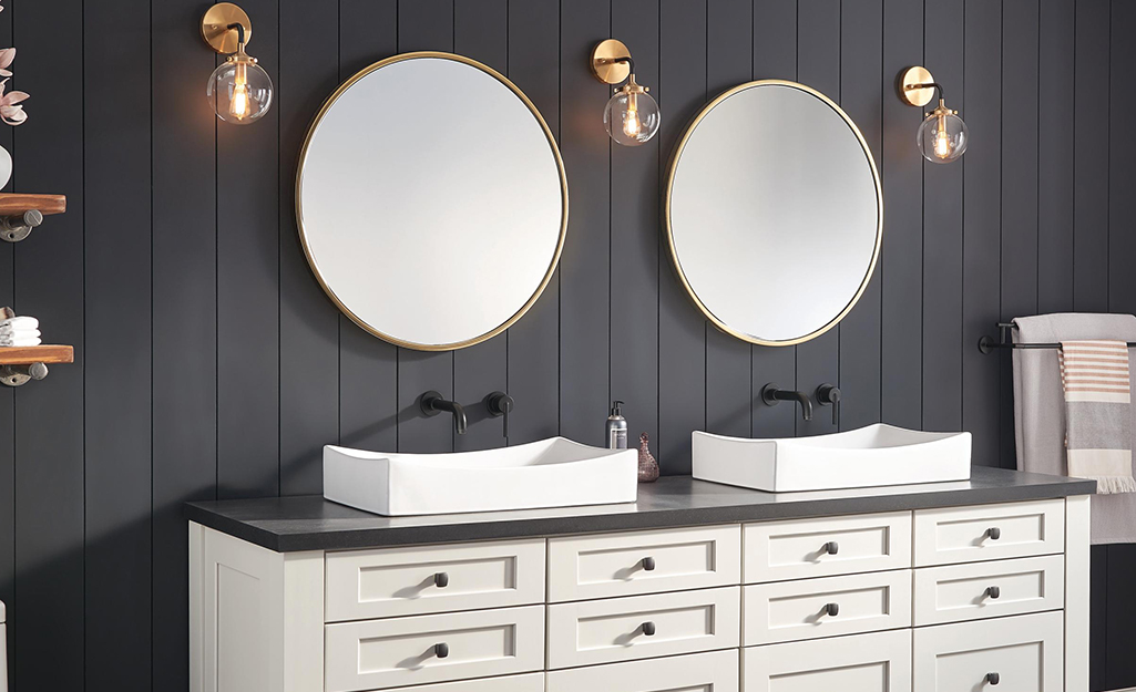 Glass globes cover the three lights installed on the sides of two circular mirrors above a dual vanity against a gray wall.