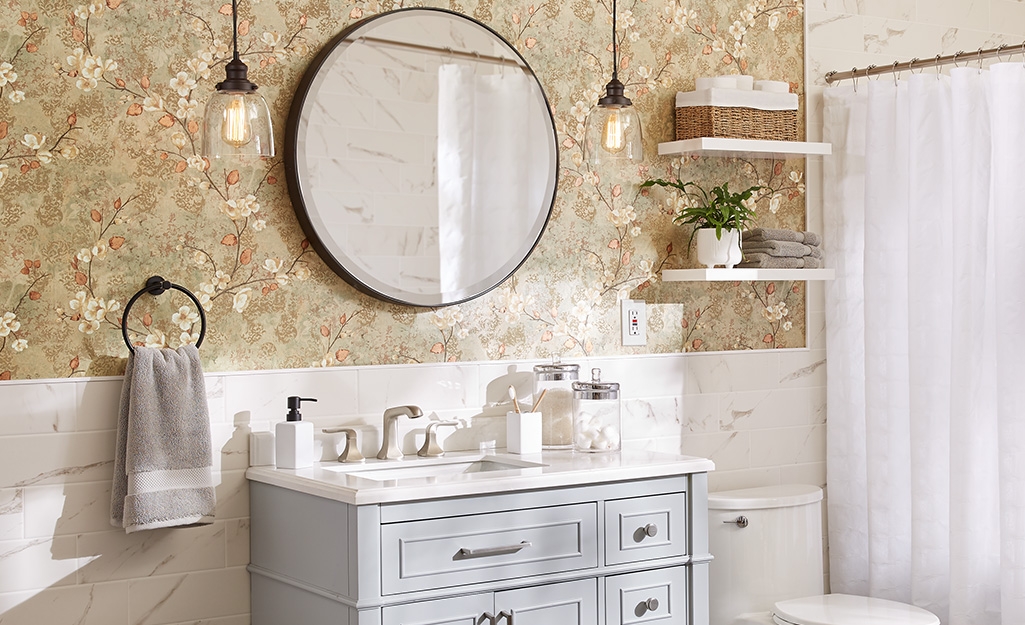 Two pendant lights hang down on each side of a circular mirror against floral wallpaper and above a bathroom vanity. 