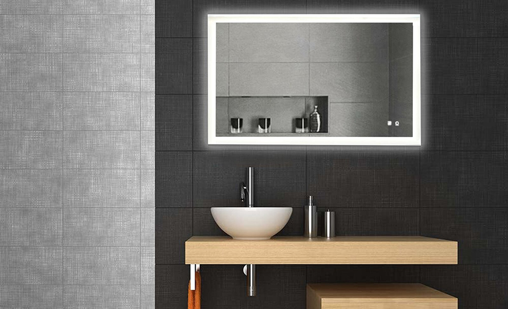 A lit mirror hanging over a wood sink in a simple black and grey bathroom.