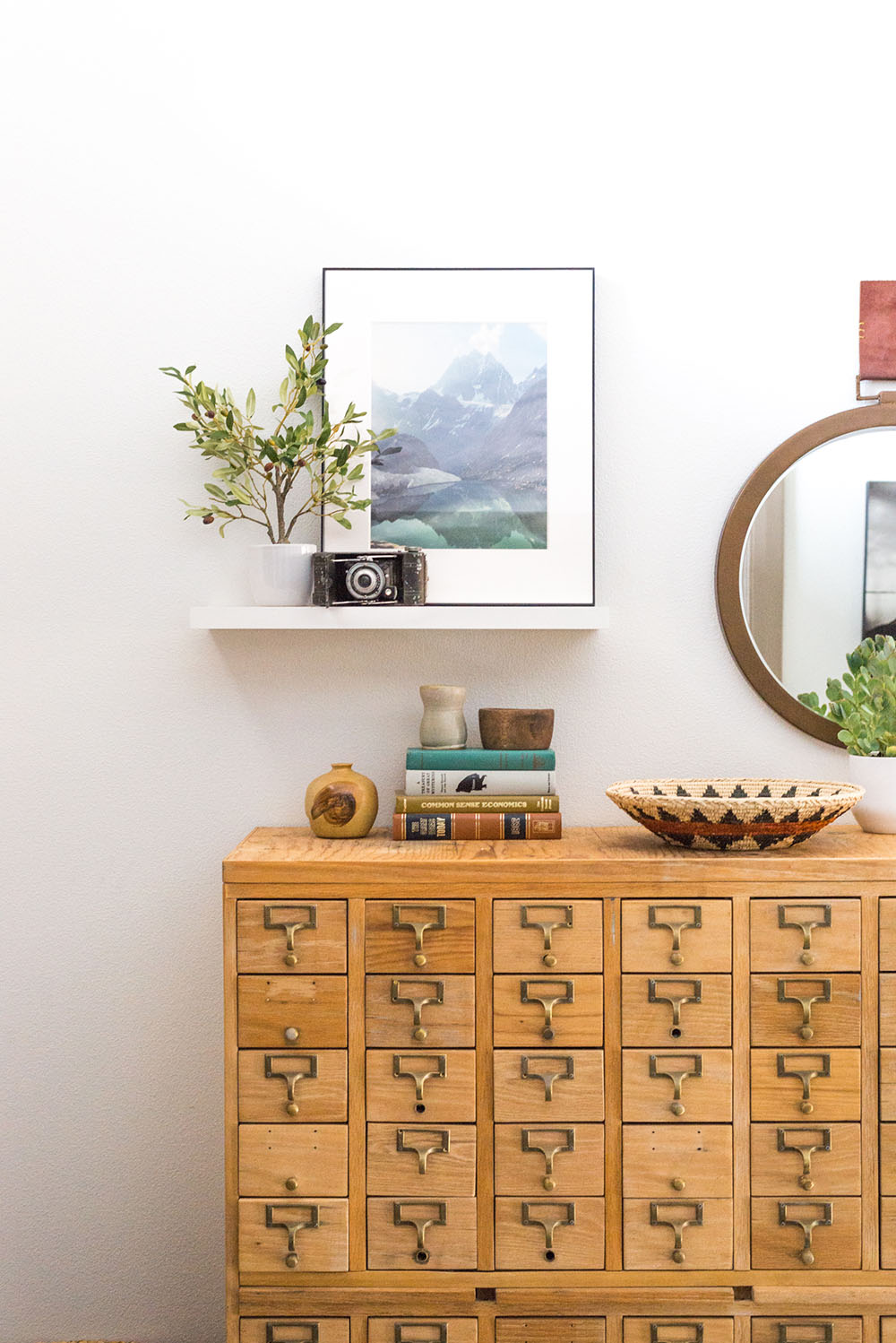 A floating shelf with a camera, plant, and picture sit above a wooden piece of furniture. A floating shelf with a camera, plant, and picture sit above a wooden piece of furniture.