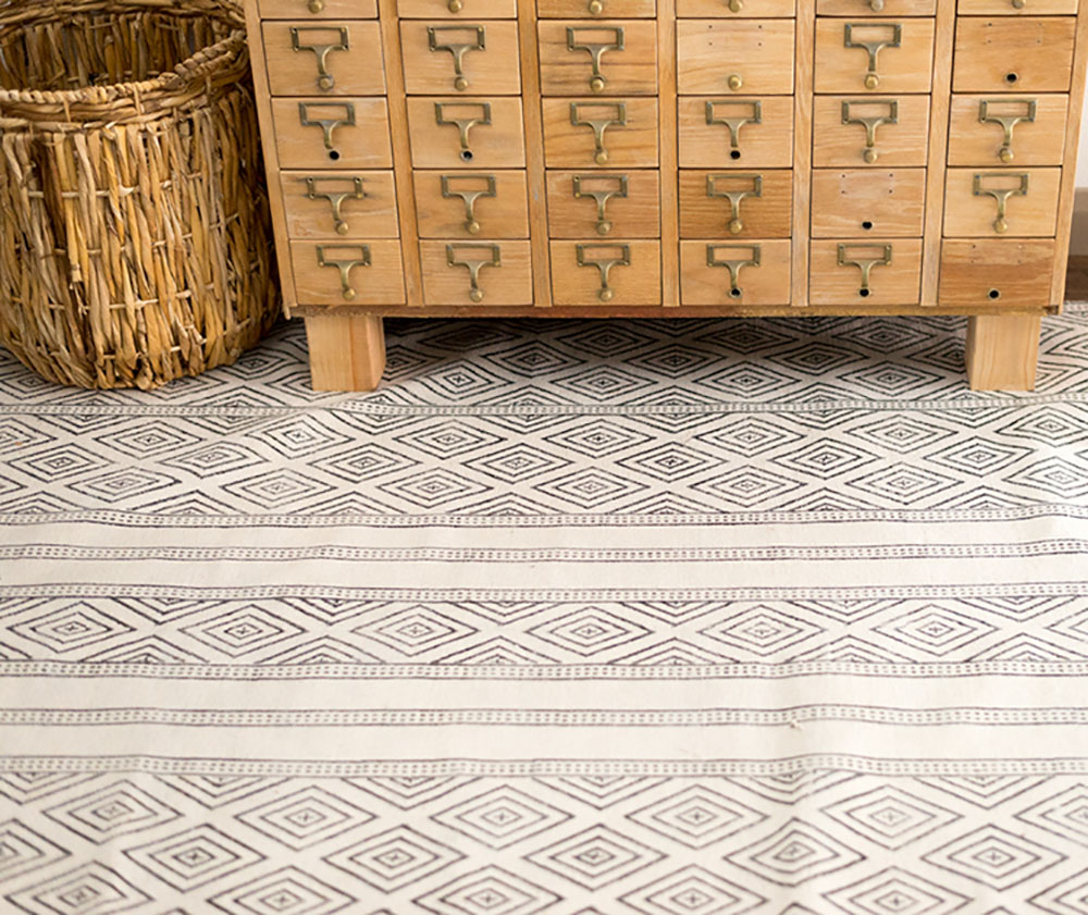 The bottom of a wooden piece of furniture and a rustic basket sit on a patterned rug. The bottom of a wooden piece of furniture and a rustic basket sit on a patterned rug.