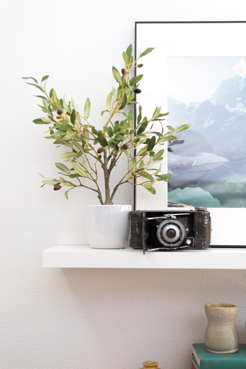 A white floating shelf with a faux olive tree, framed art, and an old camera. A white floating shelf with a faux olive tree, framed art, and an old camera.