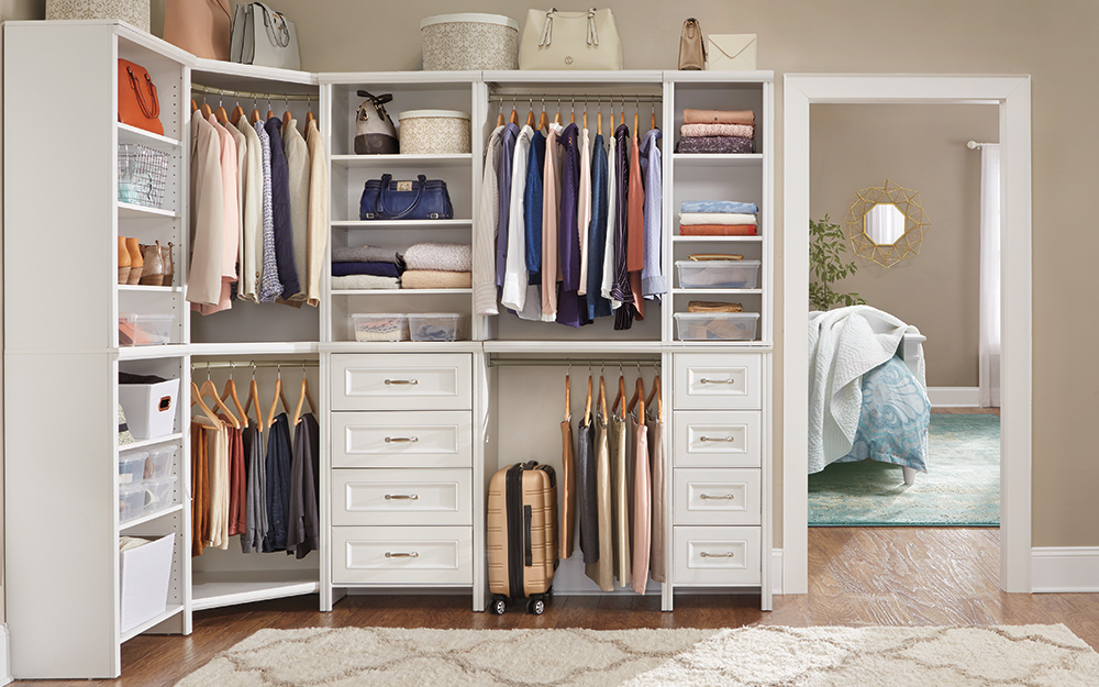 In a white walk-in closet, shirts and pants hang among shelves that hold sweaters, shoes, purses and storage bins. 