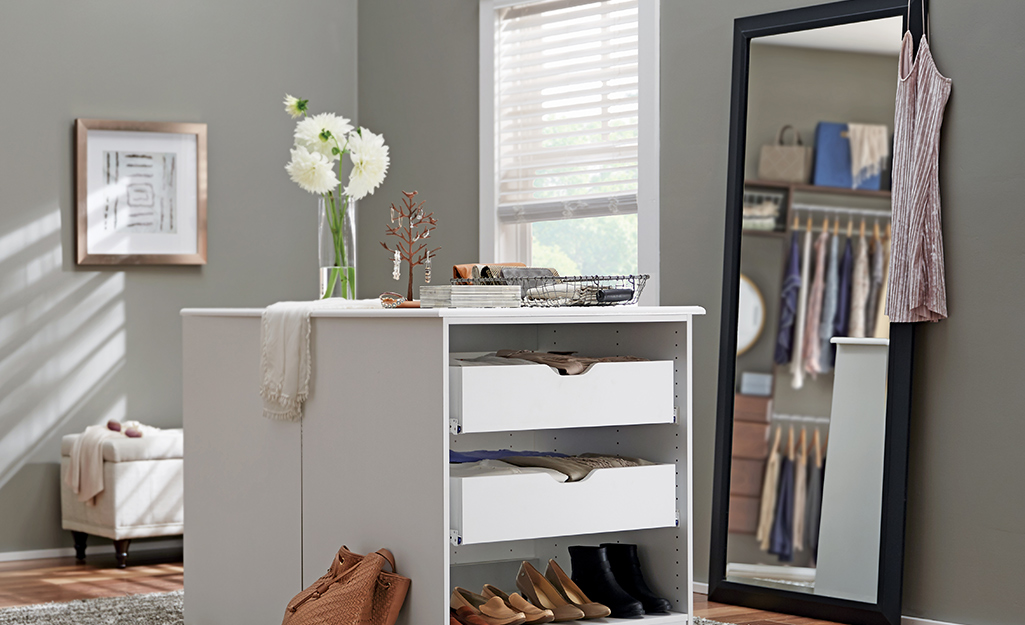 A full-length mirror against a wall and next to a storage island reflects racks of clothes in a walk-in closet.