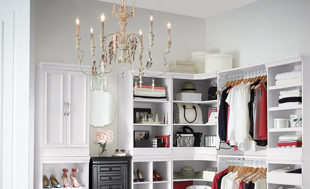 A candelabra-style chandelier hangs from the ceiling of a white walk-in closet with shelves and hanging clothes.