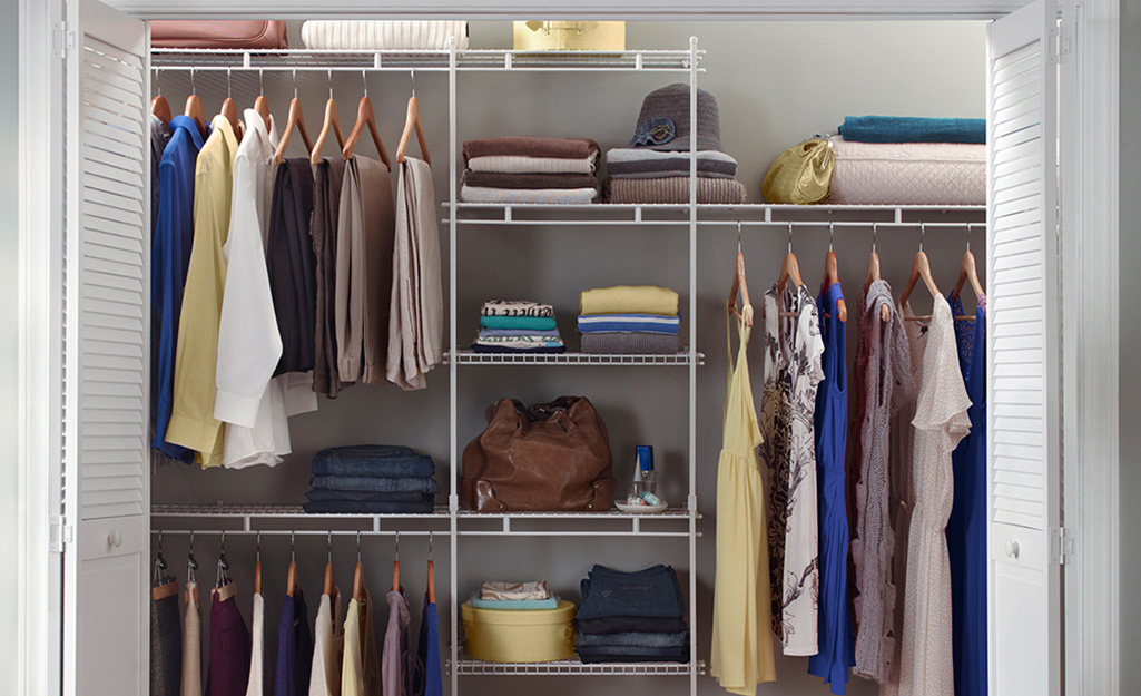 Clothes hang between white wire shelves in a closet system in a small walk-in closet.